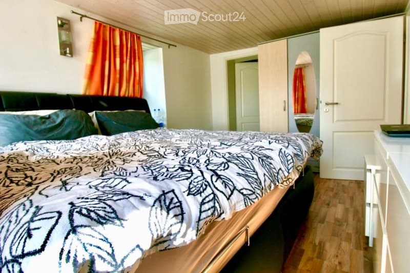 Cozy bedroom with a large bed, patterned duvet, green pillows, a black leather sofa, a white wardrobe with a mirror, and a nightstand.