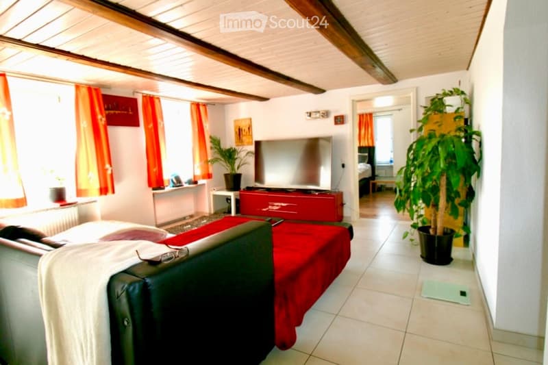 A cozy living room with a grey sofa, a red TV stand, a large flat-screen TV, a potted plant, and orange curtains. The room features exposed wooden beams on the ceiling and a tiled floor.