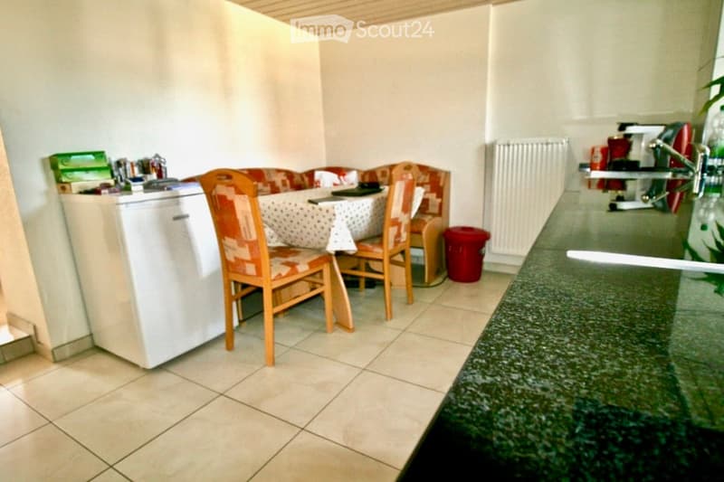 A small, furnished room with a refrigerator, a dining table with chairs, a radiator, and a trash bin. The room has tiled flooring and white walls.