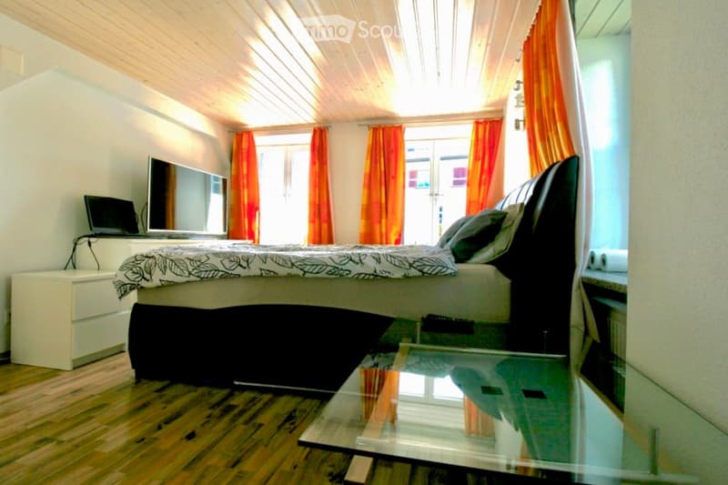 Cozy bedroom with wooden flooring, a large bed with a black and white duvet, a nightstand with a laptop, a mirror, and a glass table with a chair.