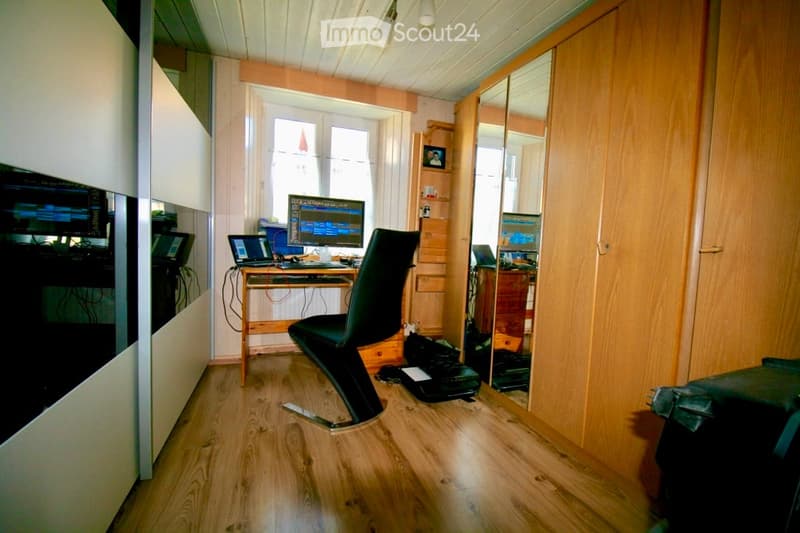 A modern, well-lit room with wooden flooring, a large window, a black leather chair, a wooden desk with a laptop and screen, and a large mirror on the wall.