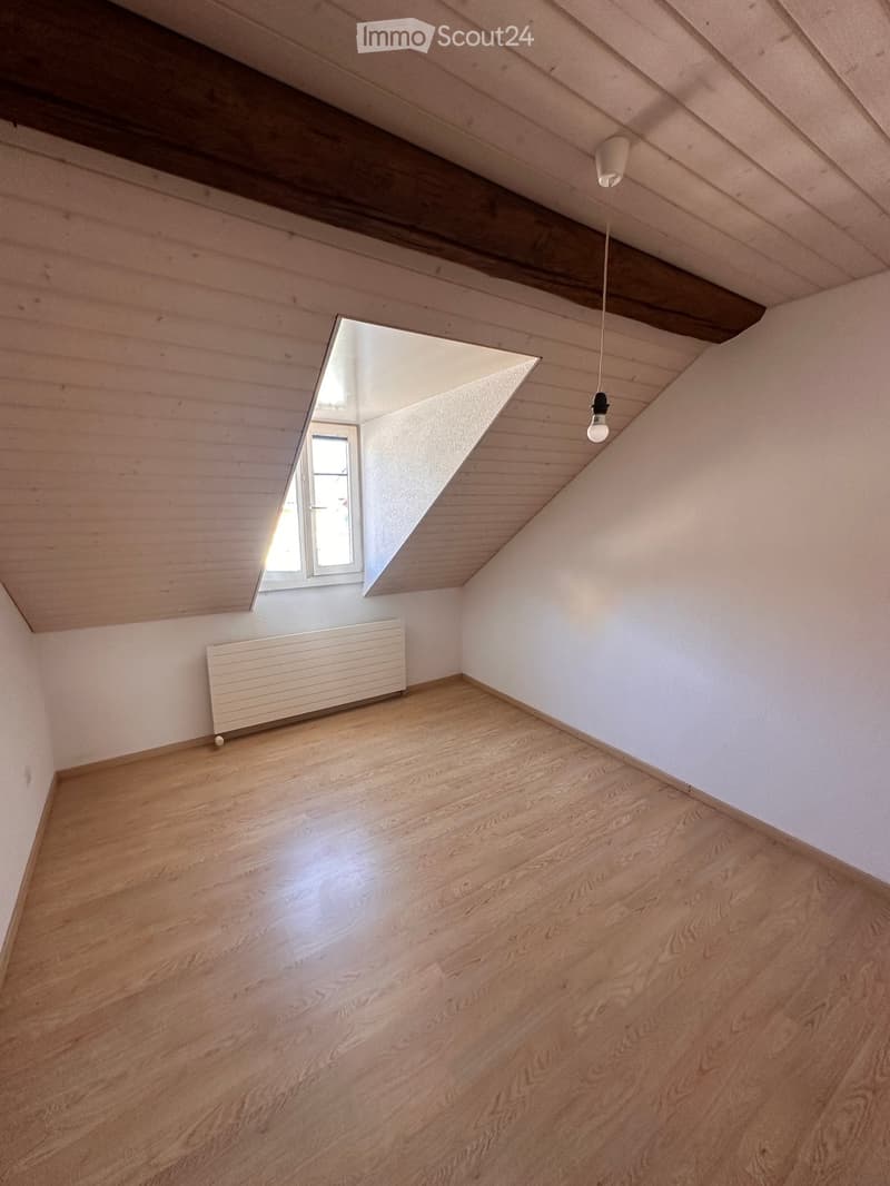 Empty room with wooden flooring, white walls, and a slanted wooden ceiling. Features a single window with white frame and a radiator on one wall.