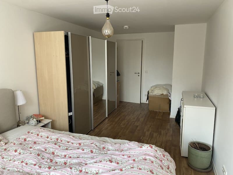 Cozy bedroom with wooden flooring, a neatly made bed with floral bedding, a nightstand with a lamp, a wardrobe, and a small refrigerator. A potted plant is placed near the refrigerator.