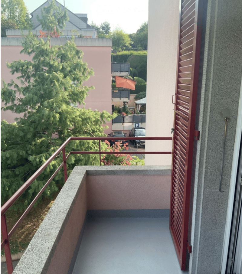 Balcony with metal railing, view of parking area and neighboring buildings, some greenery visible.