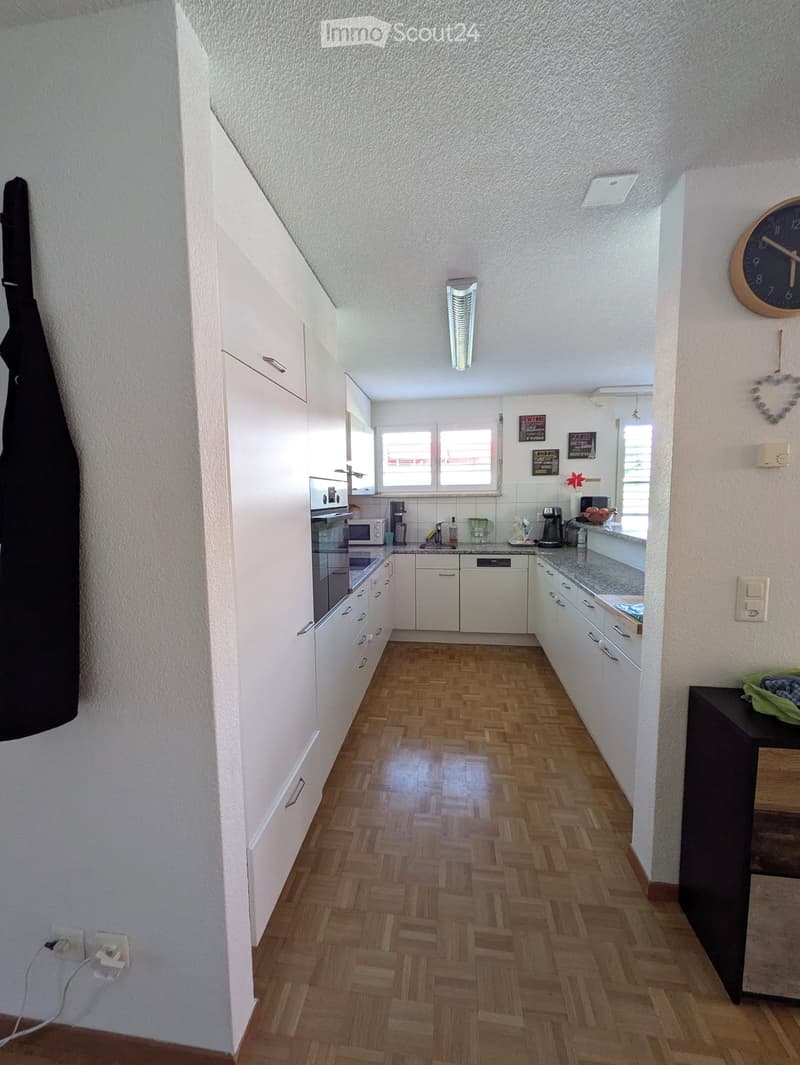 Modern kitchen with white cabinets, wooden herringbone flooring, and stainless steel appliances including an oven, microwave, and coffee maker. A window provides natural light, and a small table with a green cloth is visible on the right.