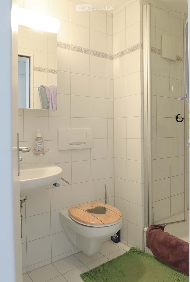 A small bathroom with white tiled walls and floor, featuring a toilet with a wooden seat cover, a sink with a mirror above it, and a glass-enclosed shower. Towels and a bath rug are visible.