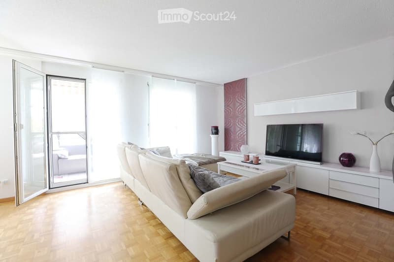Modern living room with light-colored furniture, a large sectional sofa, a coffee table, a television, and decorative items on a white cabinet.