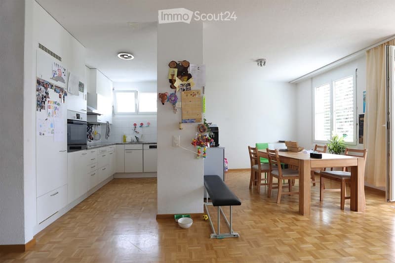 Open-plan kitchen and dining area with white cabinets, wooden flooring, and large windows. Includes a refrigerator, oven, dishwasher, sink, dining table with chairs, and a small stool.