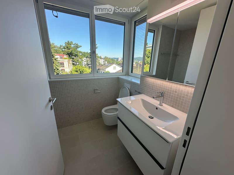 Modern bathroom with a white sink, chrome faucet, toilet, and large mirror. The room features tiled walls and a view of the city through large windows.