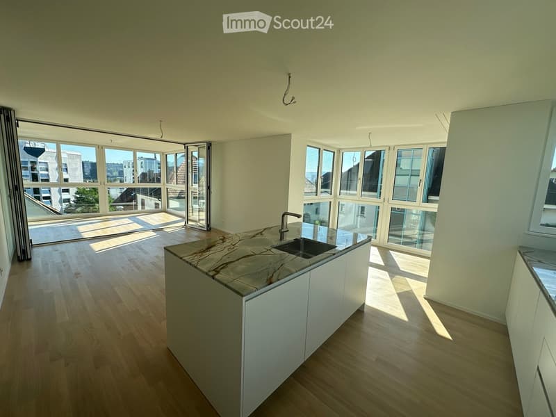 Modern kitchen with light wooden flooring, white cabinets, marble countertop, and large windows offering a view of the city.