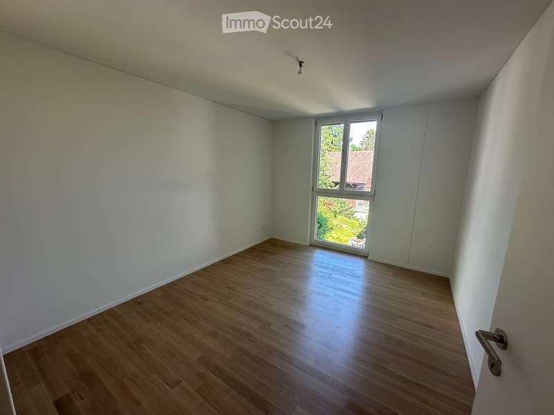 Empty room with light wooden flooring, white walls, and a large window with a view of greenery outside.