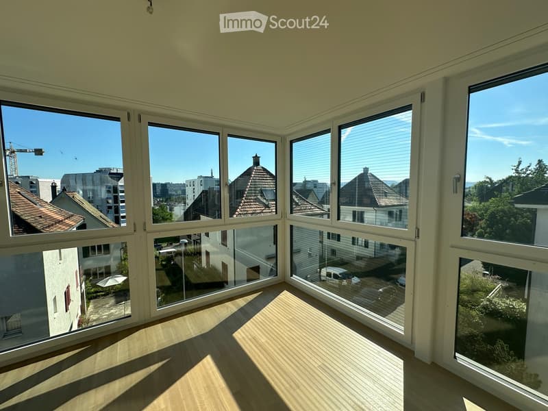 Empty room with large windows offering a view of the cityscape and surrounding buildings. Light wooden flooring and white walls.
