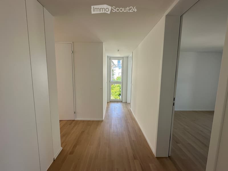 Empty hallway with wooden flooring, white walls, and a window at the end letting in natural light.