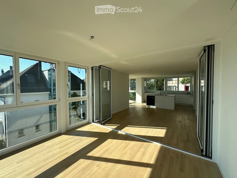 Bright, empty room with light wooden flooring and large windows, connected to a modern kitchen area with white cabinets and stainless steel appliances.