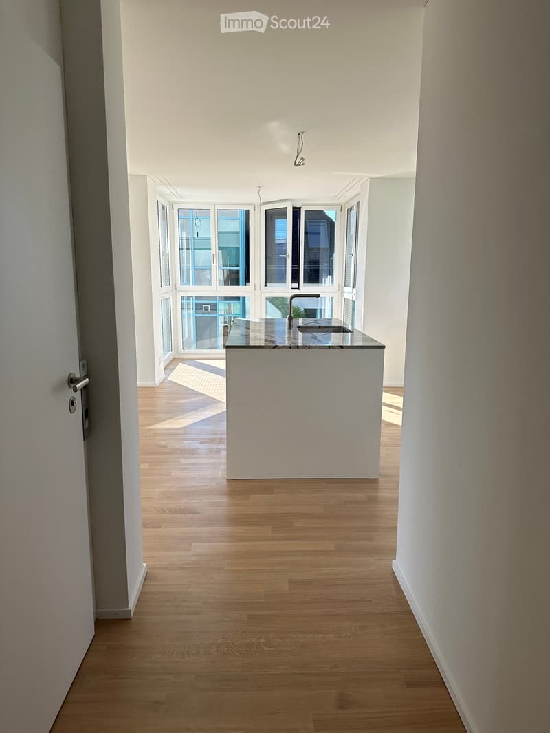Empty hallway with wooden flooring, white walls, and a small kitchen area visible at the end.
