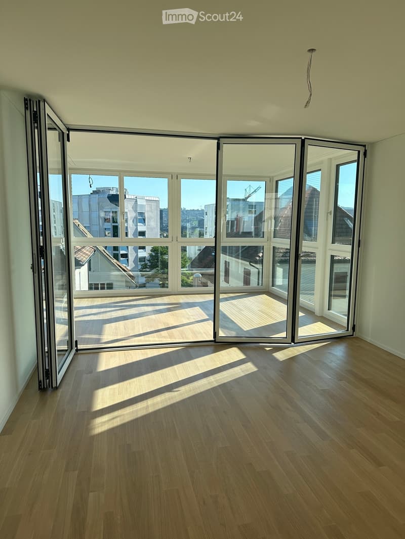 Empty room with light wooden flooring and large, fully open sliding glass doors leading to a balcony or outdoor area. The room has white walls and a modern, minimalist design.
