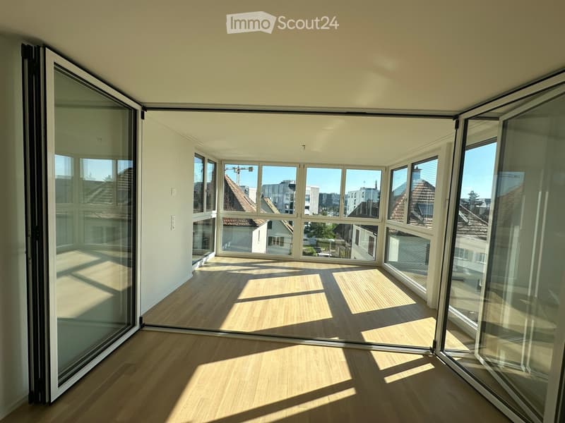 Bright, empty room with large glass windows and sliding doors, wooden flooring, and natural light.