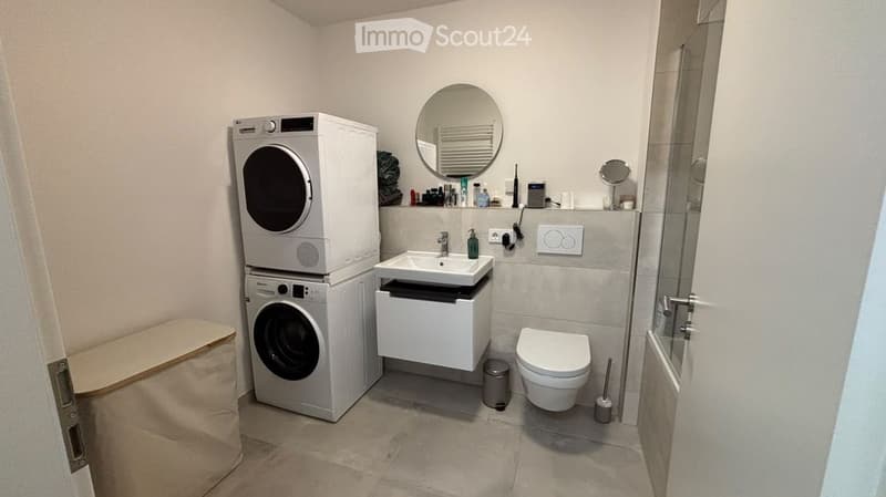 Modern bathroom with a white sink, mirror, toilet, washing machine, dryer, and a small cabinet under the sink.