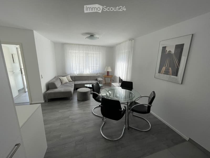 Modern living room with grey sectional sofa, glass dining table with four chairs, small round stool, side table with lamp, and large picture frame on wall.