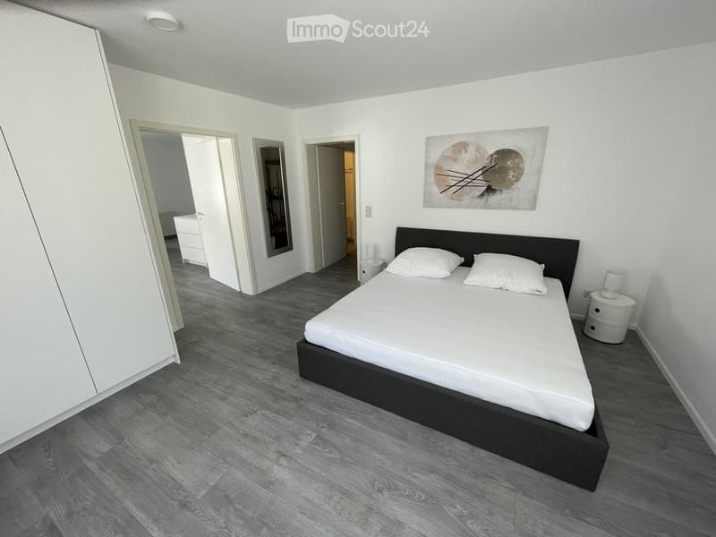 Modern bedroom with a double bed, white bedding, nightstand, mirror, and artwork on the wall. The room features light-colored walls and a dark wooden floor.
