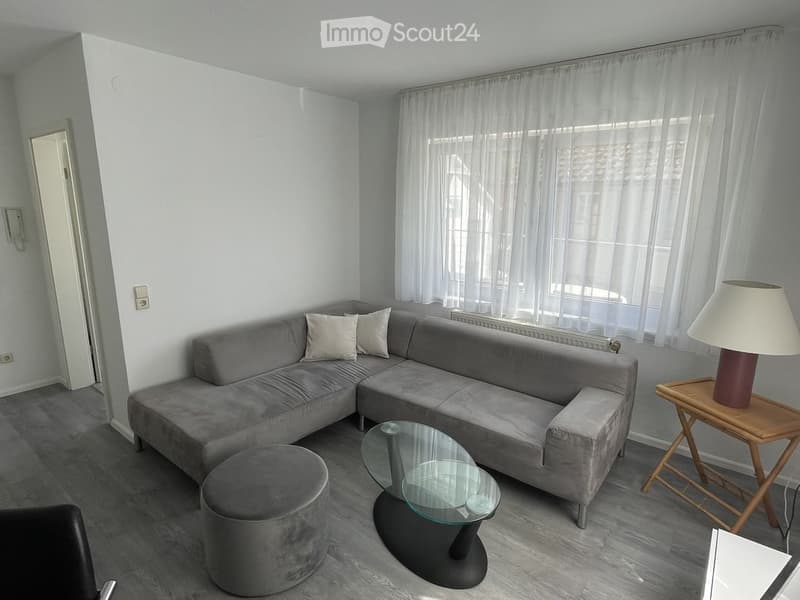 Modern living room with grey sectional sofa, ottoman, glass coffee table, side table with lamp, and large windows with white curtains.