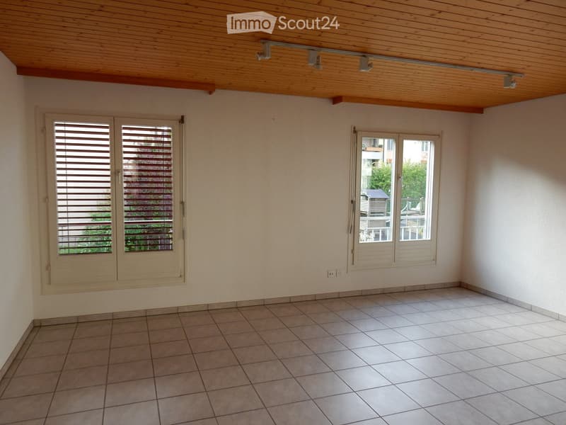 This image shows a spacious room with a wooden ceiling, white walls, and tile flooring. The room has large windows that provide natural light and views of the surrounding greenery. The room appears to be empty, with no furniture or decor visible.