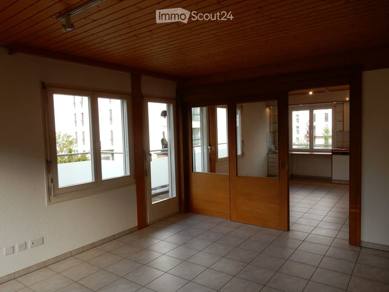 The image shows a spacious room with a wooden ceiling, large windows, and tiled flooring. The room has a wooden partition wall that separates it from another area, possibly a kitchen or dining room. The overall atmosphere appears to be warm and inviting.