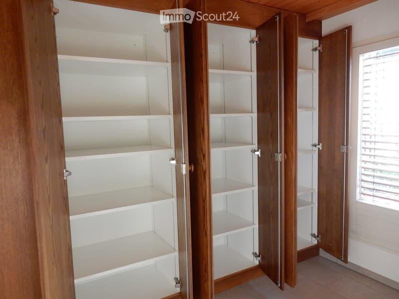 The image shows a bedroom with a built-in wardrobe. The wardrobe has multiple shelves and compartments, providing ample storage space. The walls are made of wood paneling, giving the room a warm and cozy feel. The room also has a window, allowing natural
