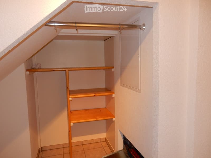 The image shows a small closet space with wooden shelves and a hanging rod. The closet has a white wall and tile flooring.