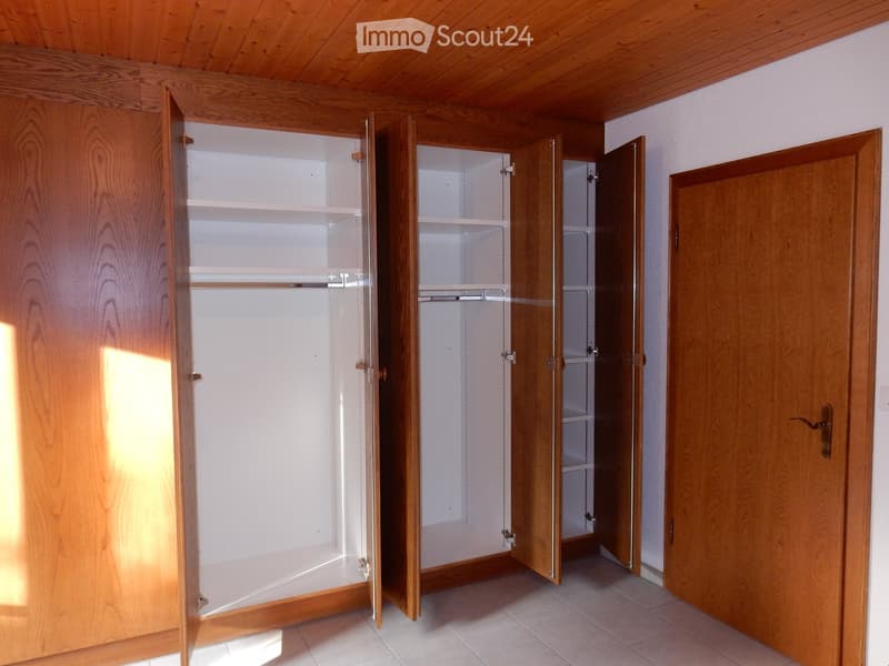 The image shows a bedroom with a wooden ceiling and walls. There are three built-in closets or wardrobes with white shelves and doors. The floor appears to be tiled. The room has a simple, functional design.