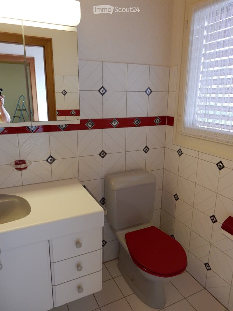 Tiled walls with red and white pattern, white vanity with drawers, toilet, window