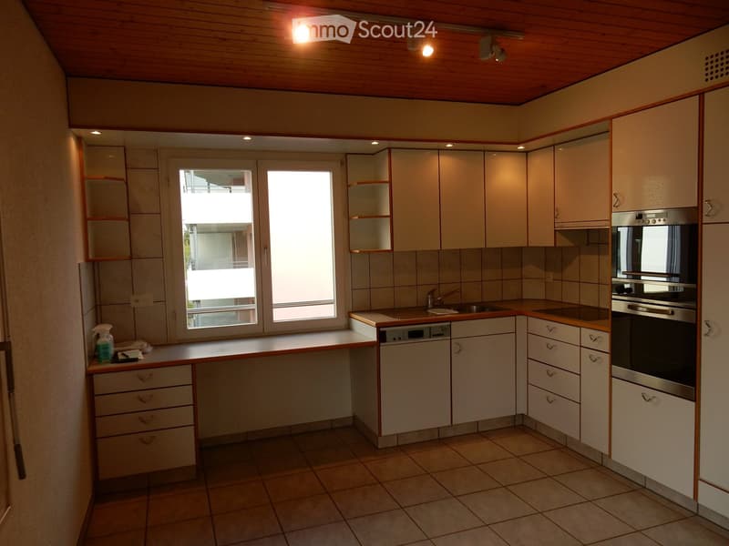 Spacious kitchen with wooden cabinets, tile flooring, built-in appliances including a dishwasher, and large windows providing natural light.