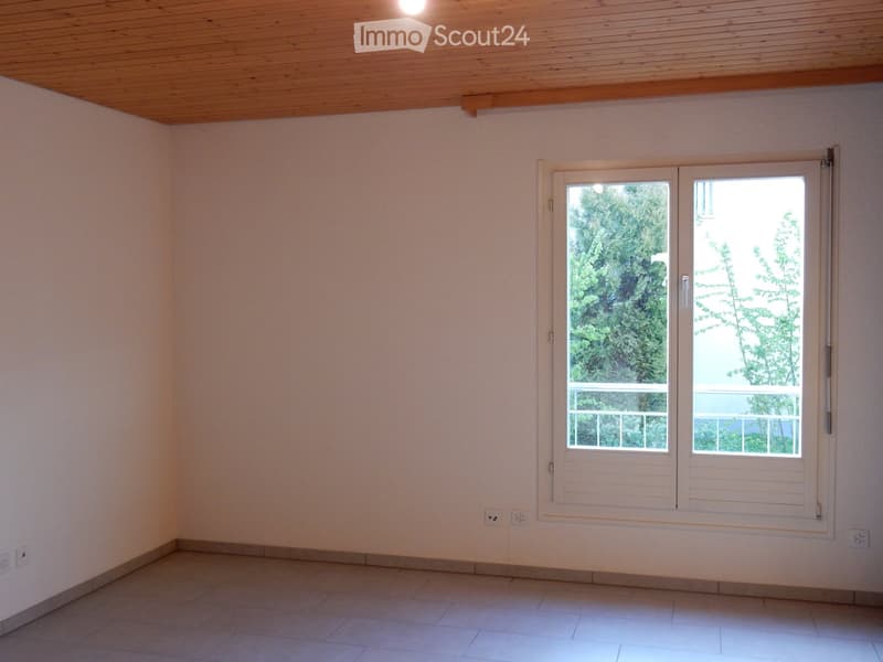 Spacious room with wooden ceiling, white walls, and large windows overlooking a balcony or terrace with a view of the surrounding greenery.
