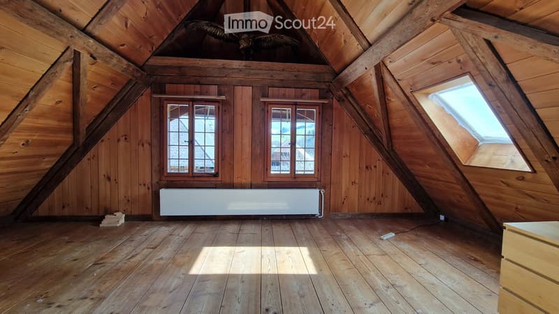 Empty attic space with wooden walls and floor, exposed wooden beams, two windows, and a radiator. No furniture or equipment present.