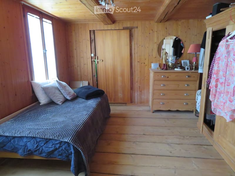 Cozy wooden bedroom with a single bed, pillows, a blanket, a wardrobe, a mirror, a lamp, and a small table with drawers.