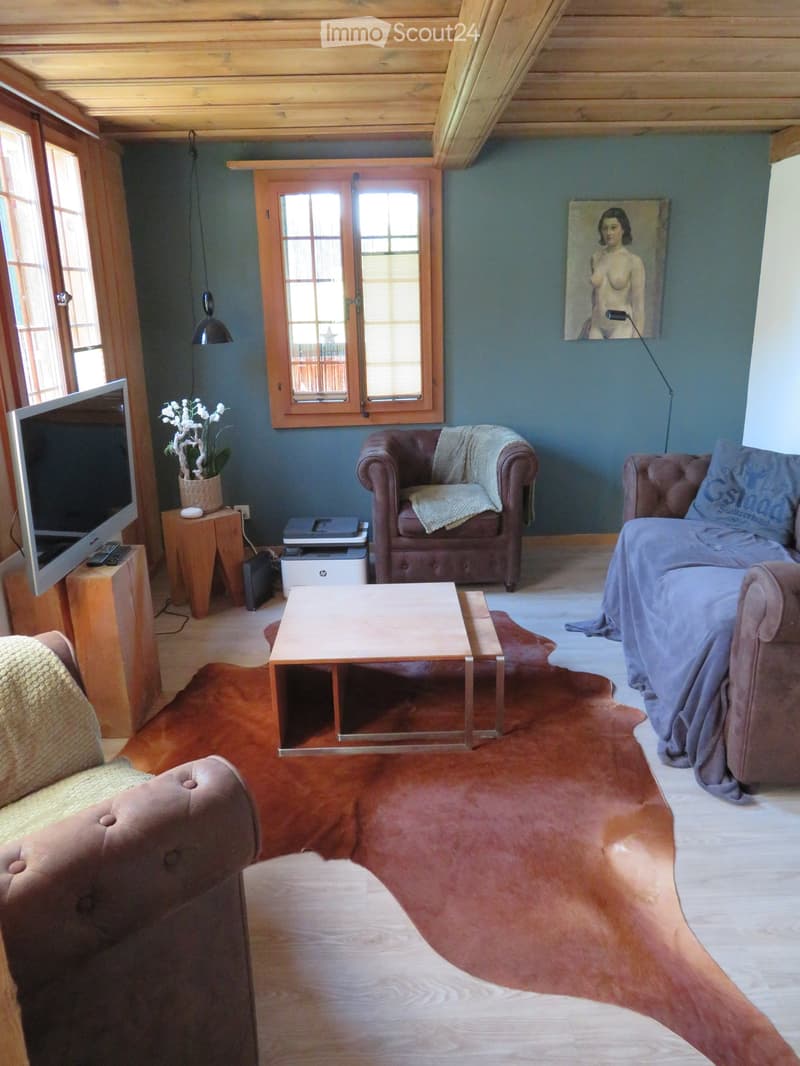 Living room with wooden ceiling, large windows, grey sofa, coffee table, armchairs, TV, and a painting on the wall.