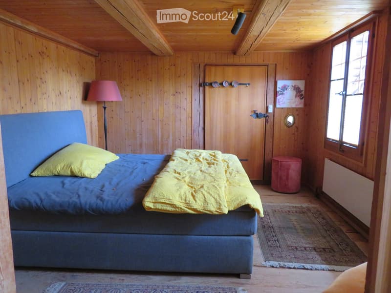 Cozy wooden bedroom with a blue bed, yellow pillows and blanket, a red stool, a floor lamp, and a picture frame on the wall.