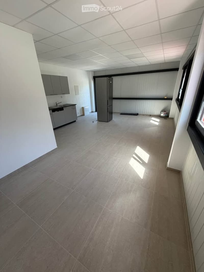 Empty room with tiled floor, white walls, and basic kitchen area with cabinets and a refrigerator. No furniture or personal items present.