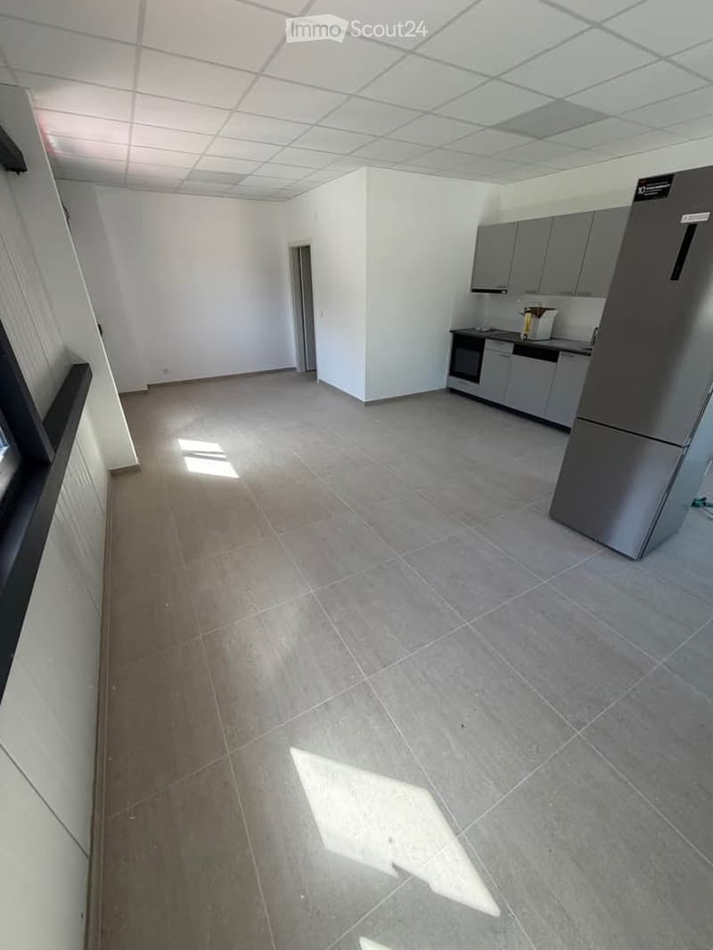 Empty room with grey tiled floor, white walls, and a small kitchen area with cabinets and appliances.