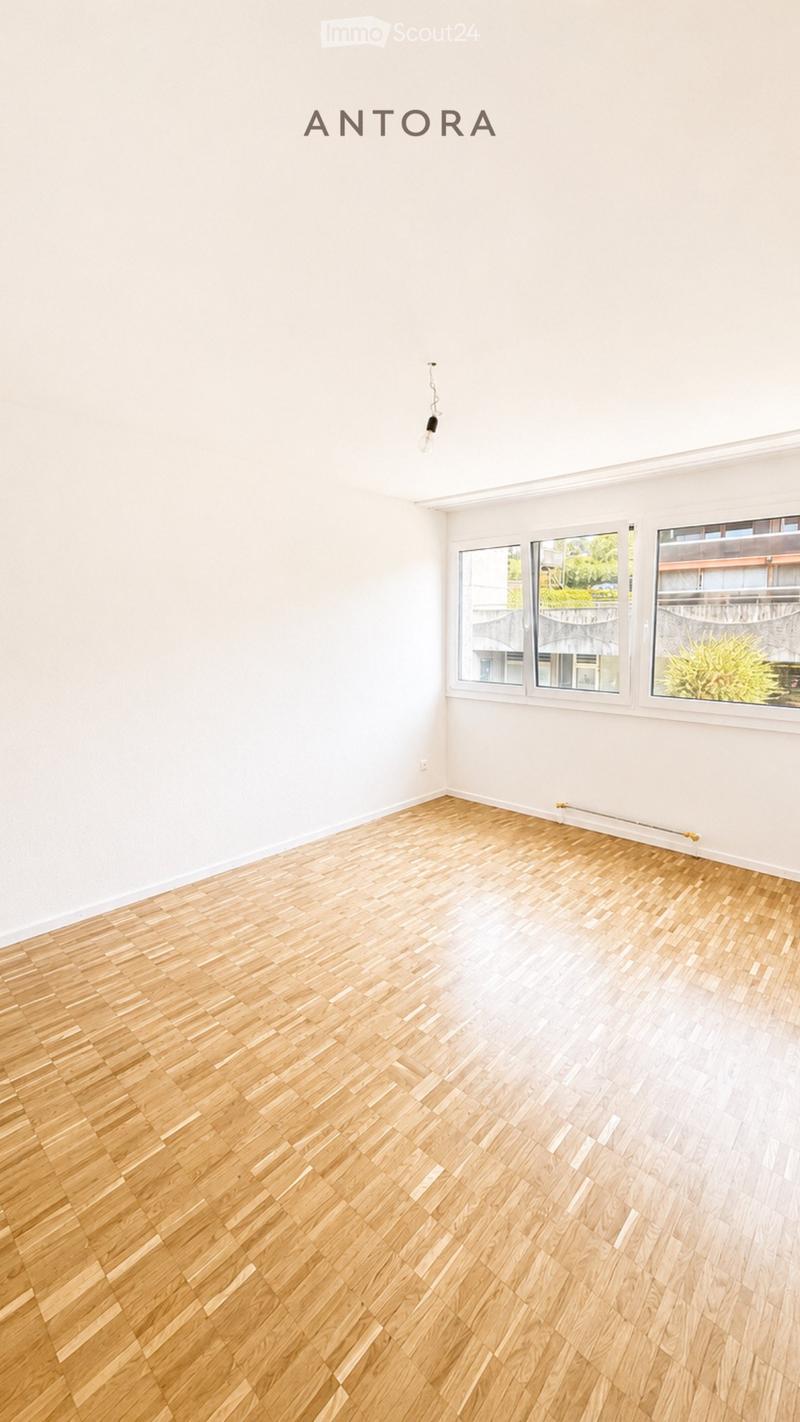 Empty room with light wooden flooring and white walls, featuring large windows with a view of the outside.