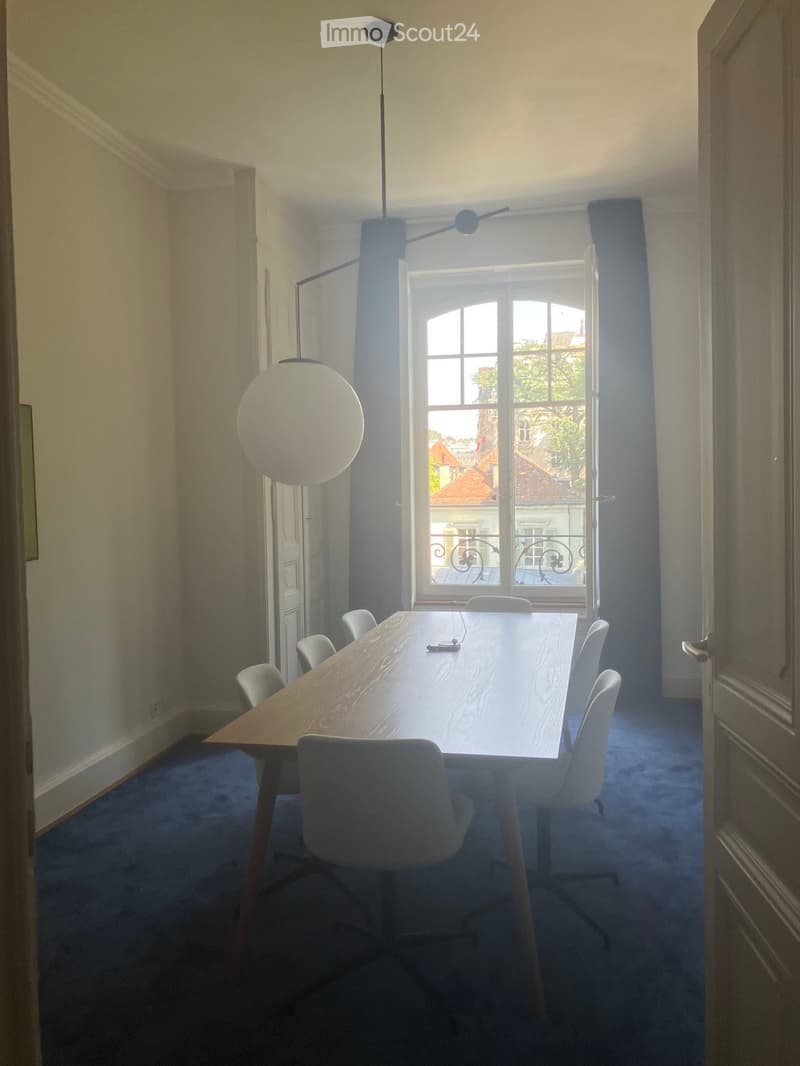 A modern, well-lit room with a long wooden table surrounded by white chairs, large windows with a view, and contemporary lighting fixtures.