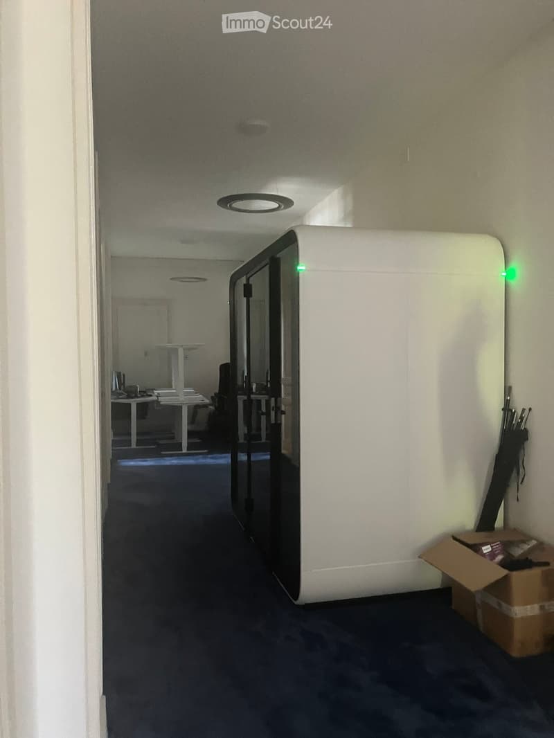 A modern hallway with white walls, dark carpeted floor, and recessed lighting. A glass door leads to an office area with desks and chairs visible in the background. An open cardboard box is on the floor near the wall.