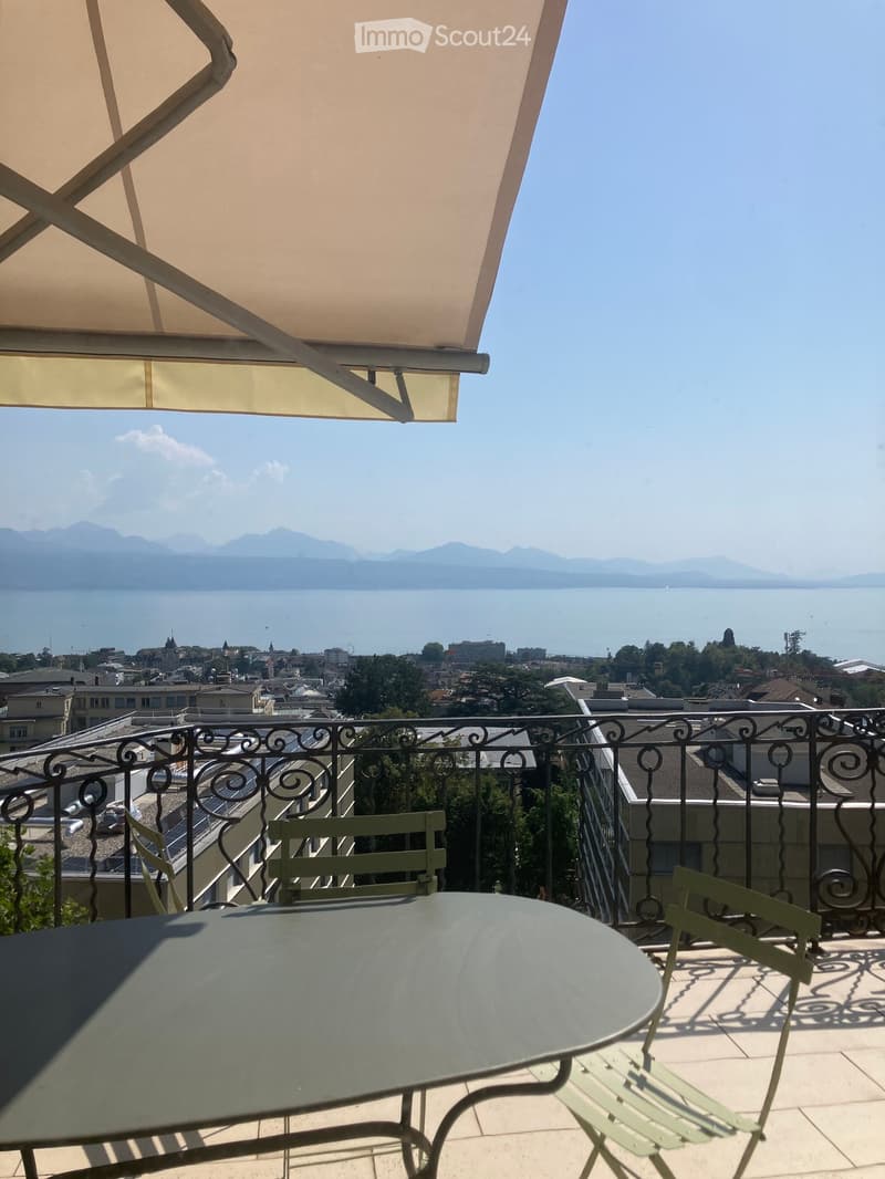 Balcony with a round glass table and two green chairs, overlooking a scenic lake and distant mountains.