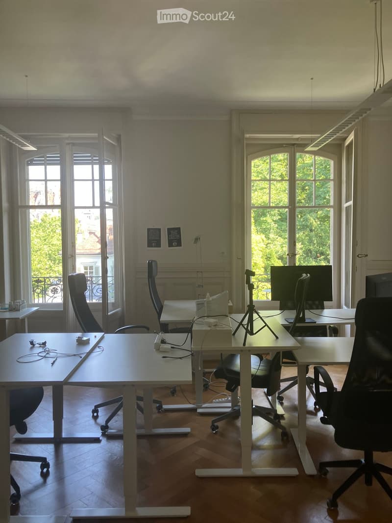 Bright, spacious room with wooden flooring, large windows, multiple desks, chairs, and a tripod with a camera setup.