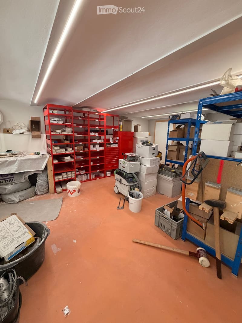 Empty storage room with red and blue shelving units, various boxes, tools, and equipment scattered around. The floor is painted orange, and the walls are white.