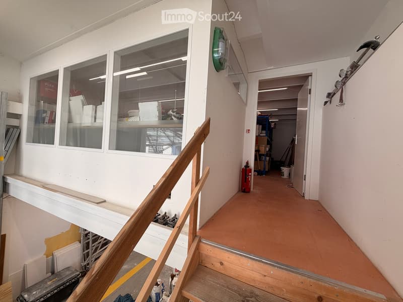 Empty hallway with wooden staircase, white walls, and large windows, showing construction materials and tools.