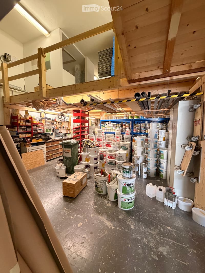 An empty workshop or storage area with exposed wooden beams, concrete floor, and scattered paint buckets, tools, and storage shelves.