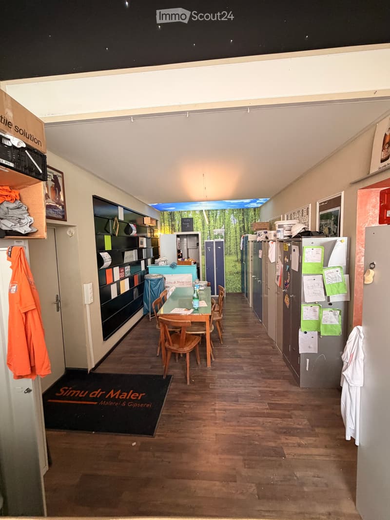 A well-lit room with wooden flooring, a table with chairs, shelves, lockers, and a refrigerator. The walls are painted in neutral tones, and there is a large window with a scenic view.