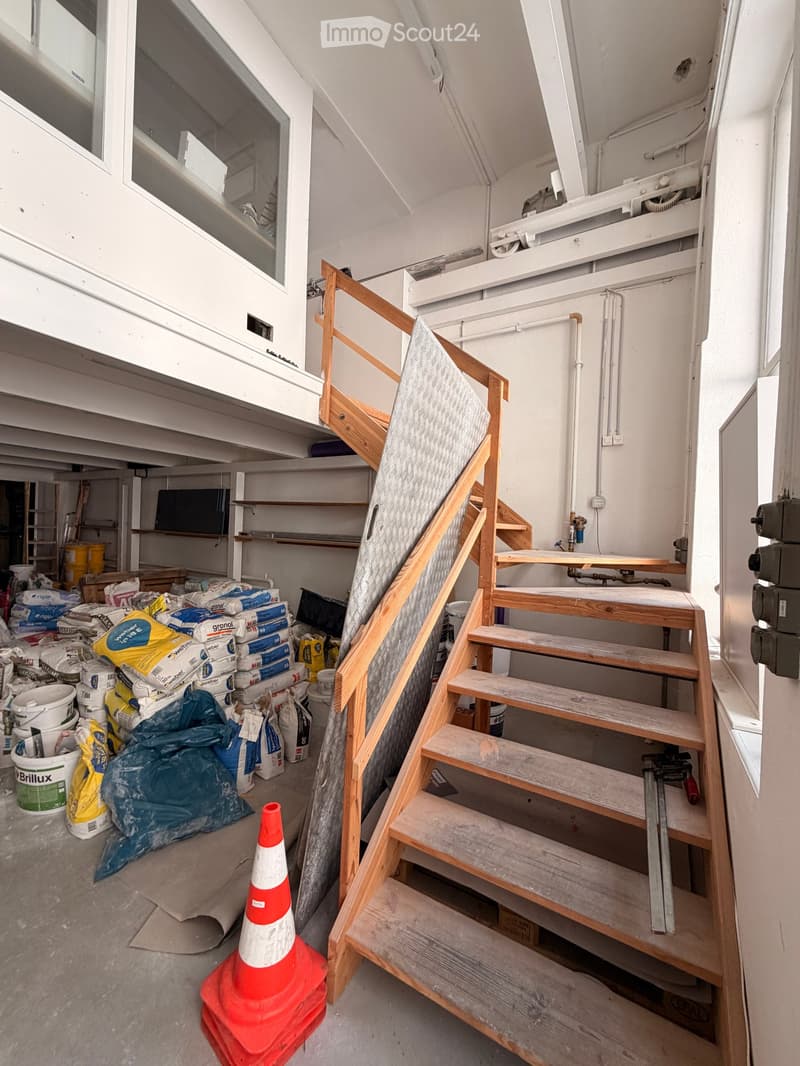 Empty hallway with a wooden staircase leading upwards, construction materials and tools scattered on the floor, and a traffic cone placed near the stairs.