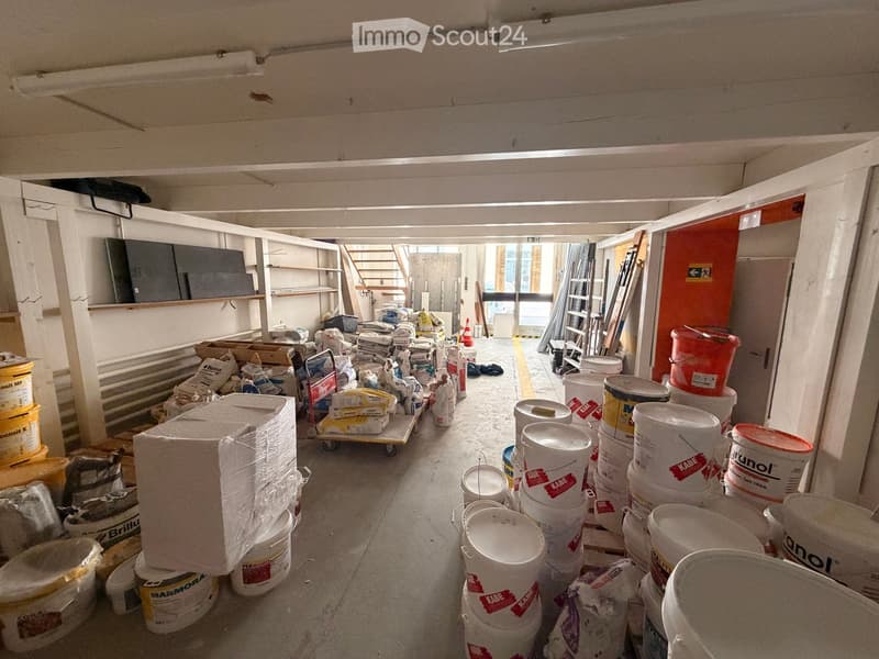 A cluttered storage area with various construction materials, including paint buckets, boxes, and tools, arranged along the walls and floor.